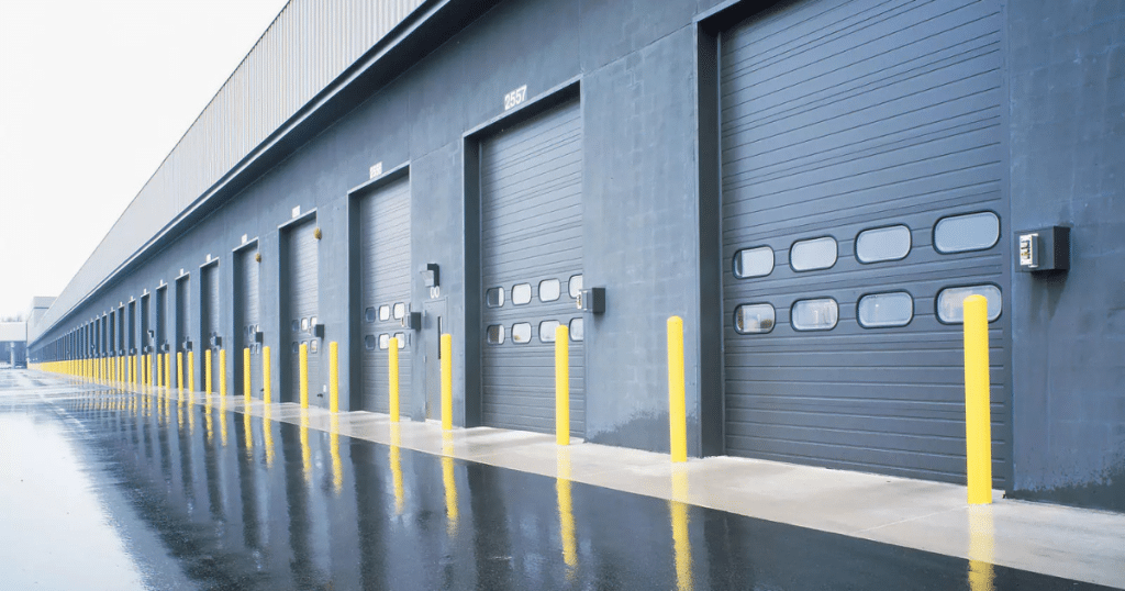 A row of large gray industrial doors with yellow bollards in front.